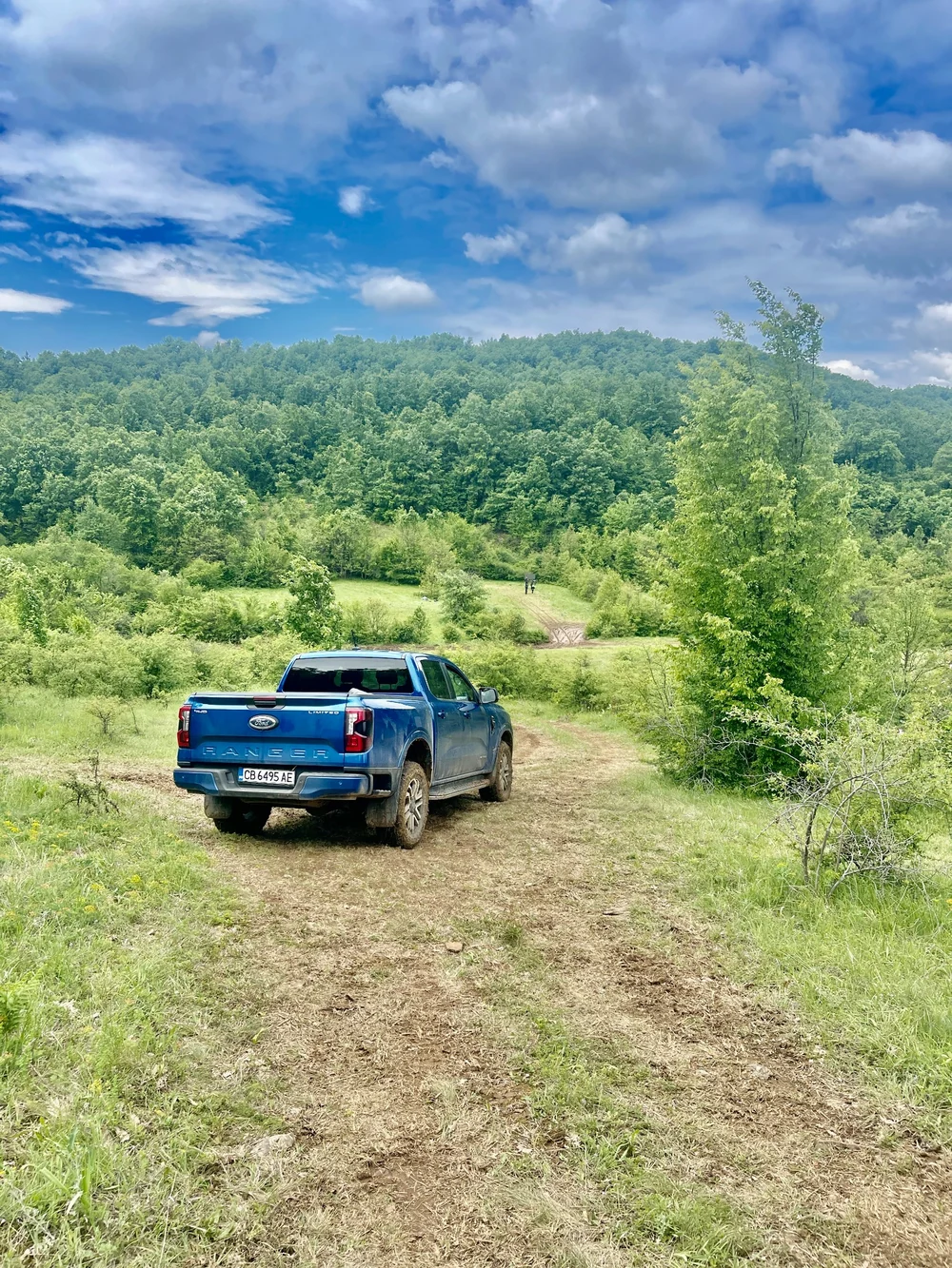 A blue four-wheel-drive pickup on an unpaved track through wooded hills